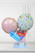 A Back to School balloon bouquet gift set featuring a light blue balloon with colorful school bus illustrations and 'Back to School' text, a metallic rose gold foil balloon, a blue striped napkin, chocolate bars, and pink tissue paper in a white box, set against a white paneled wall background.
