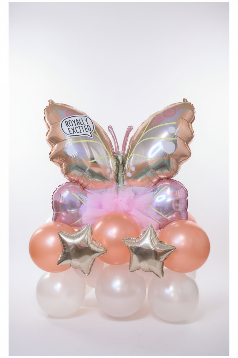 A decorative balloon bouquet shaped like a butterfly with iridescent rose-gold wings, pink tulle, peach balloons, white pearl balloons, and two metallic gold star balloons against a glittery gold background.