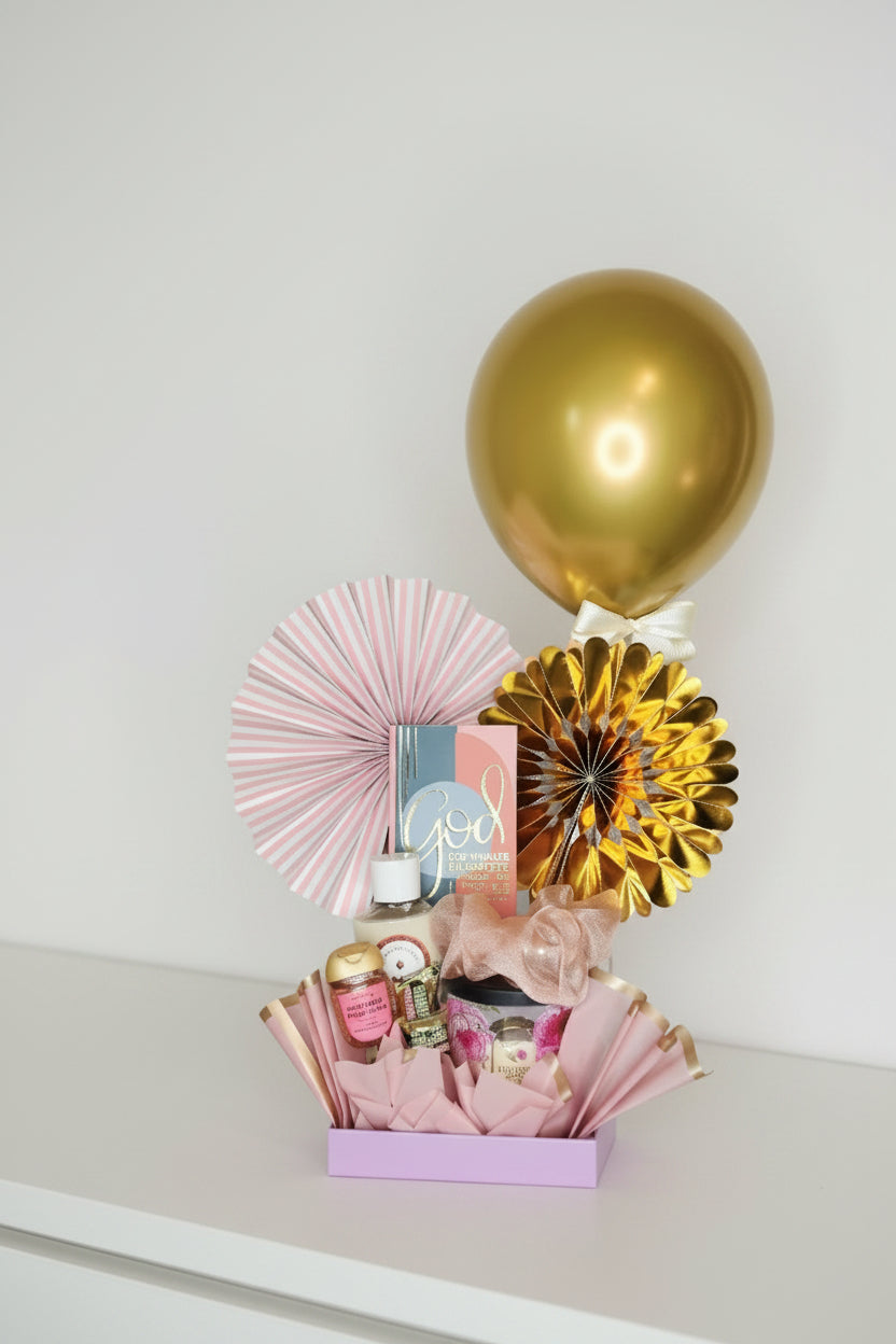 A baby shower celebration gift basket filled with pink tissue paper, small bottles, and party accessories, featuring a large gold balloon, a pink paper fan, a gold metallic paper fan, and a pink box with a gold 'B' ornament, set against a plain light gray background.